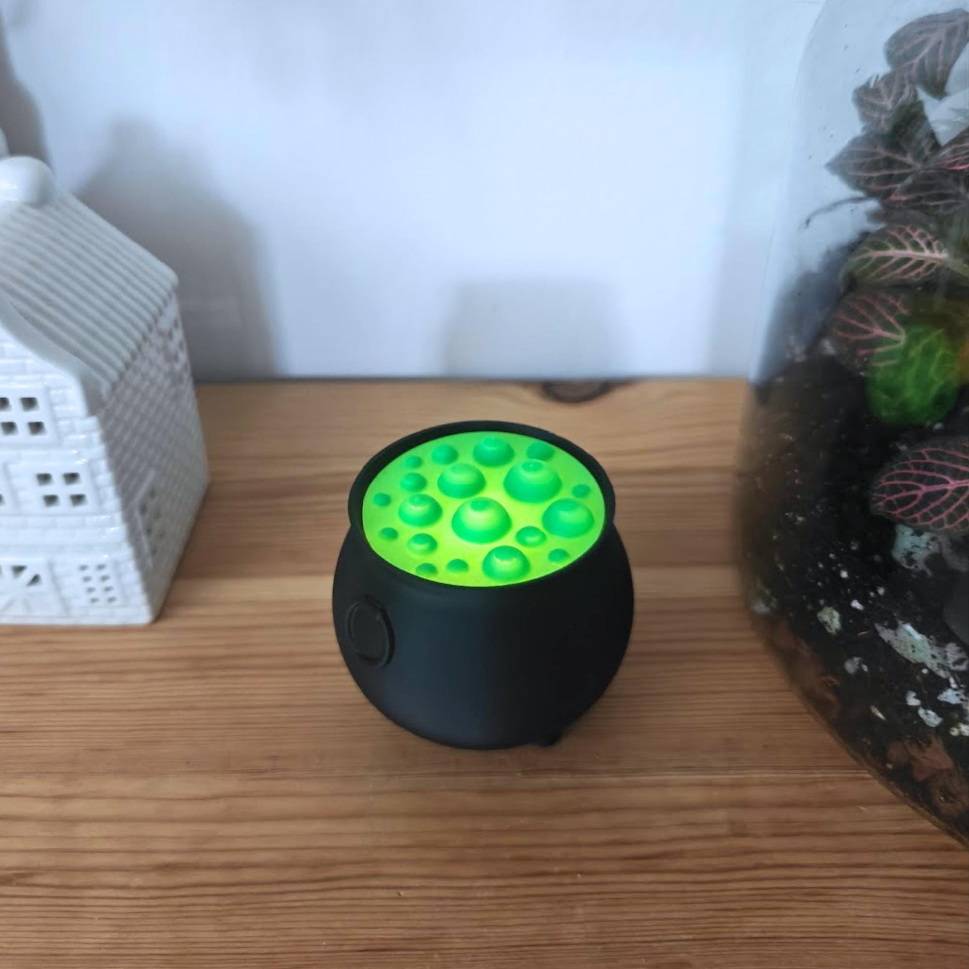 Close-up of black cauldron with green bubble lid emitting a soft glow, surrounded by decorations.