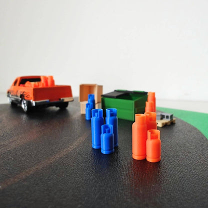 Miniature 1:64 scale scene featuring blue and orange 3D-printed gas bottles arranged near a green dumpster, wooden crate, and pickup truck on a black diorama surface.