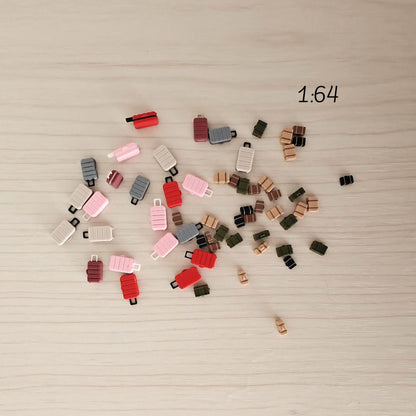 A scattered mix of 1:64 scale mini suitcases and trunks in red, pink, green, and brown tones, spread out on a wooden surface.