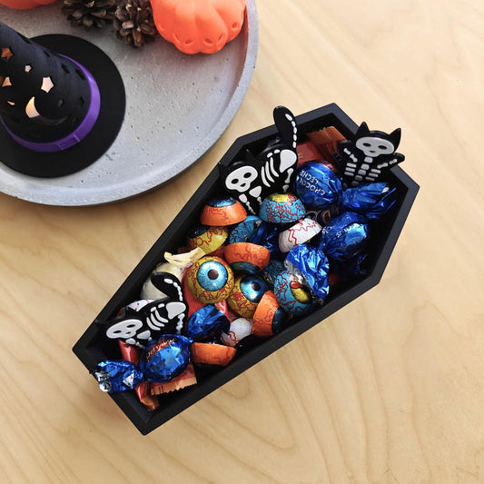 Black 3D-printed coffin-shaped candy bowl filled with colorful Halloween chocolates and mini skeleton decorations, placed on a wooden table with pumpkins