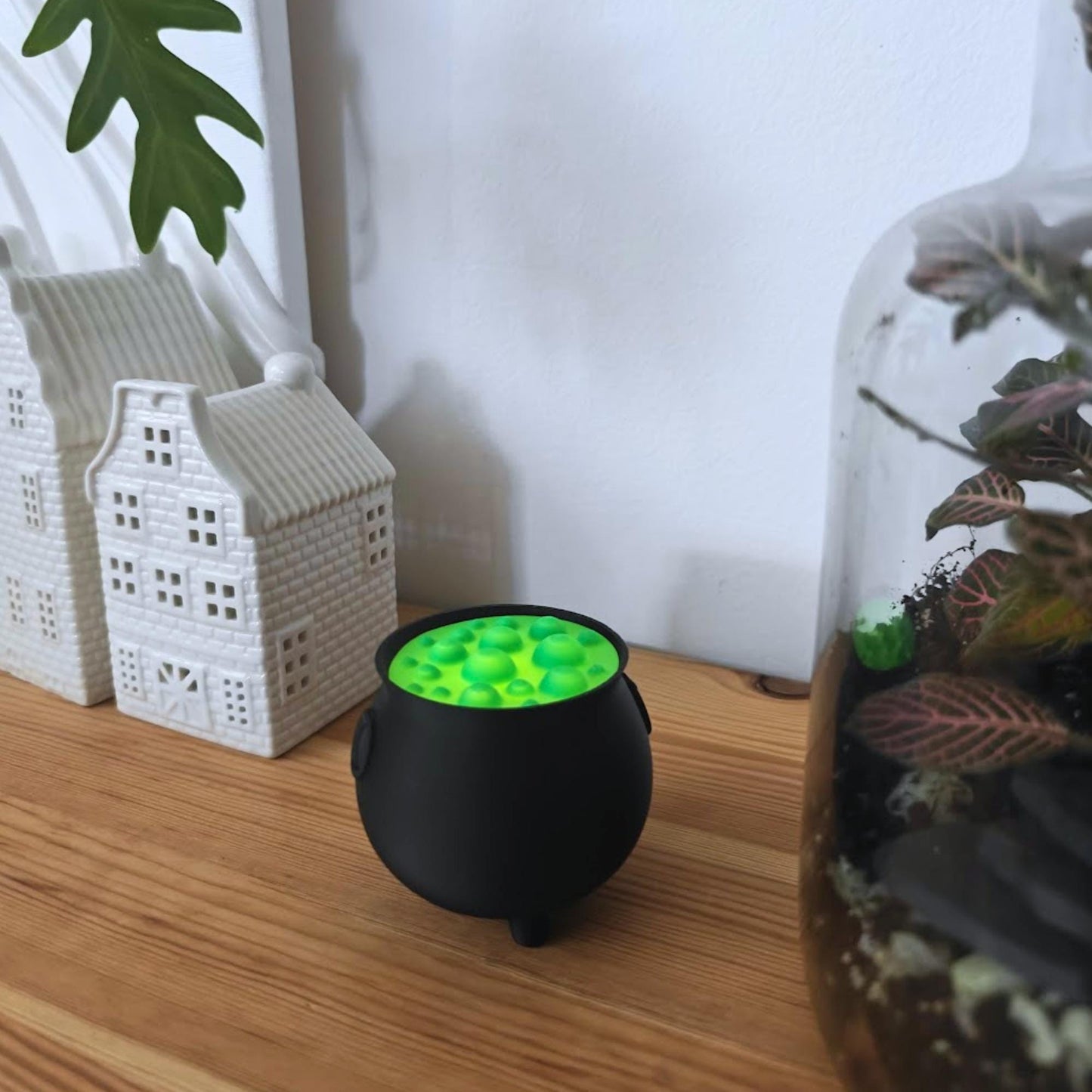 Cauldron tealight holder on wooden shelf beside white miniature houses and plants, showing bright green glow from top.