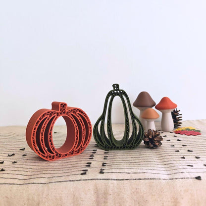 Close-up of orange and green 3D-printed pumpkins and gourds with small decorative leaves on a beige woven cloth.