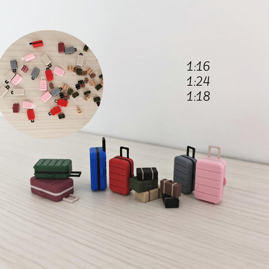 Various 3D-printed miniature suitcases and travel trunks in multiple colors and sizes, arranged in a circle on a light wood surface. Labels indicate scales 1:16, 1:24, and 1:18.