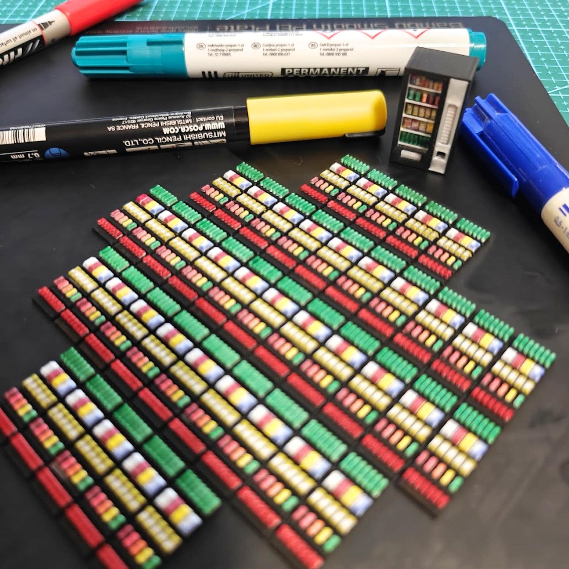 Rows of 3D-printed miniature vending machines in red, blue, green, and black lined up on a cutting mat, showing painting and detailing process.