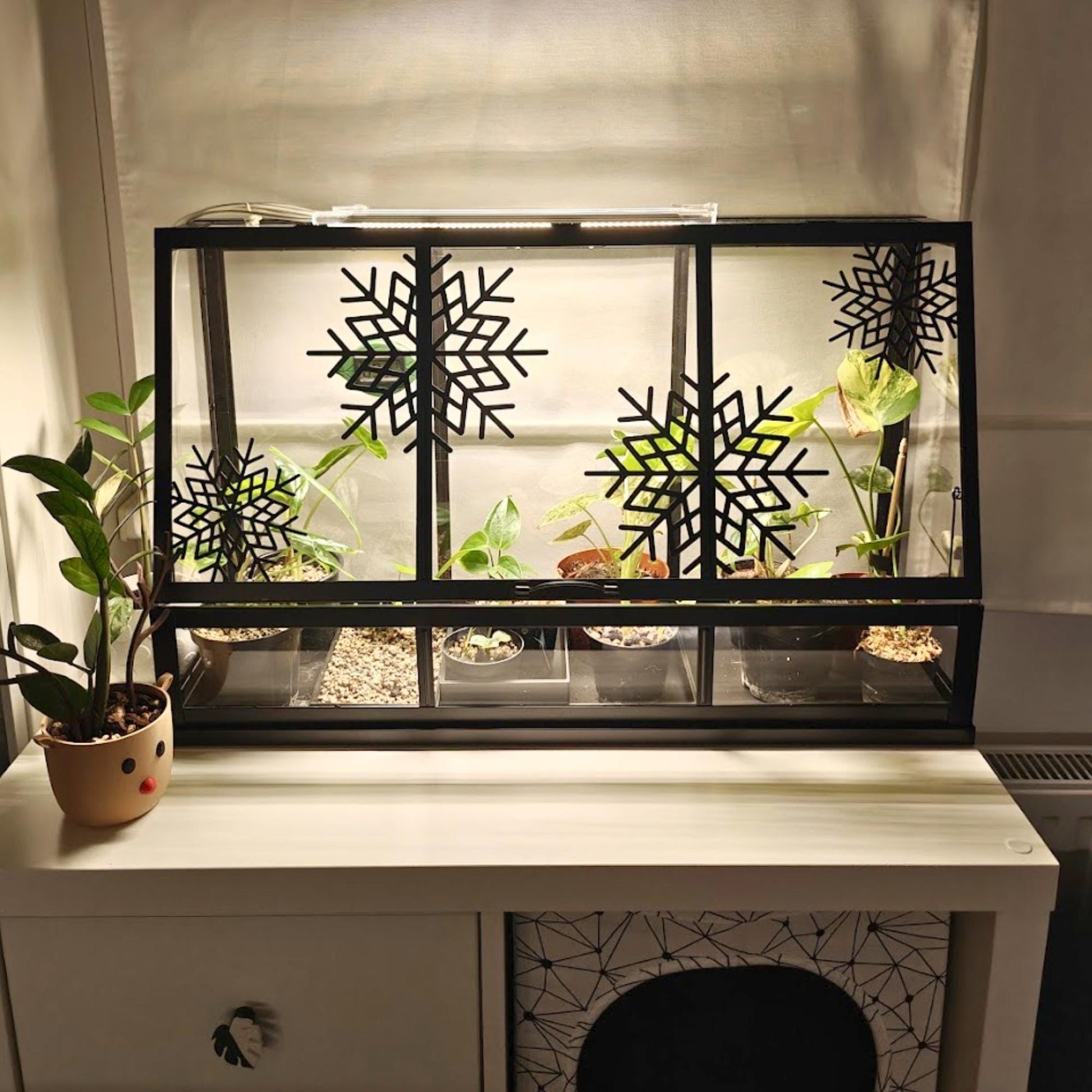 Black Äkerbar greenhouse with snowflake decorations, placed on white dresser near potted plant.