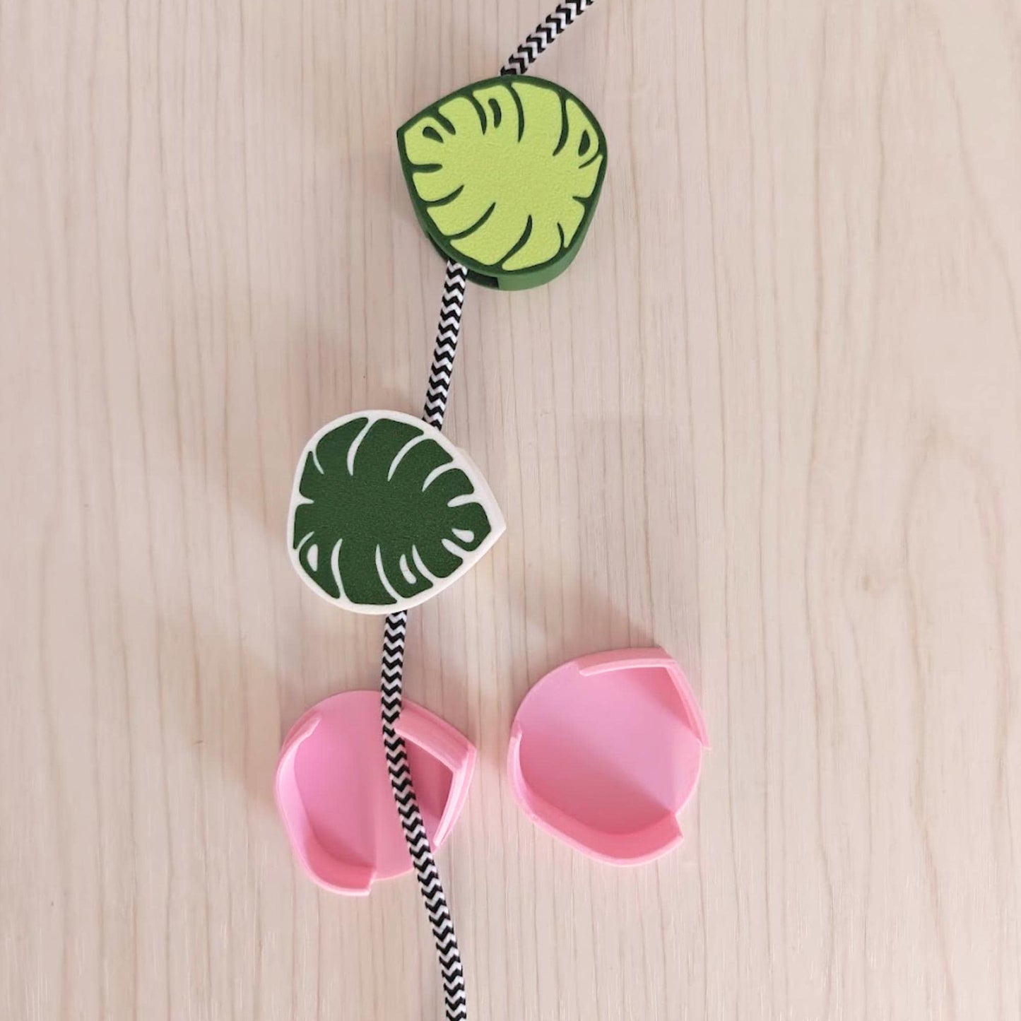 A decorative cord organiser featuring a black and white striped cord with green and white leaf-shaped clips and two pink cable clips.