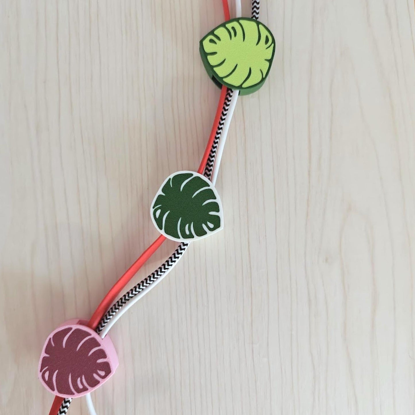 Cable clips with a leaf design in green and pink. The clips are holding patterned cords.