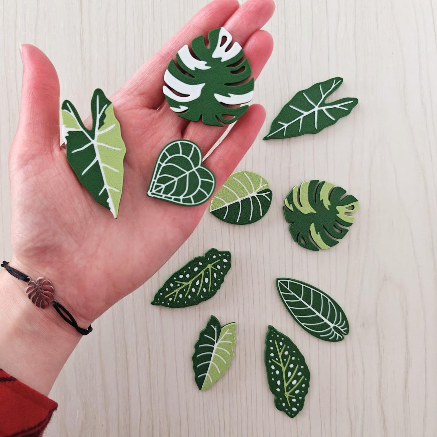 Set of 10 green leaf-shaped fridge magnets including monstera, alocasia, begonia and anthurium designs arranged in a palm for scale