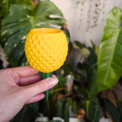 D printed pineapple watering spike held in hand in front of indoor houseplants