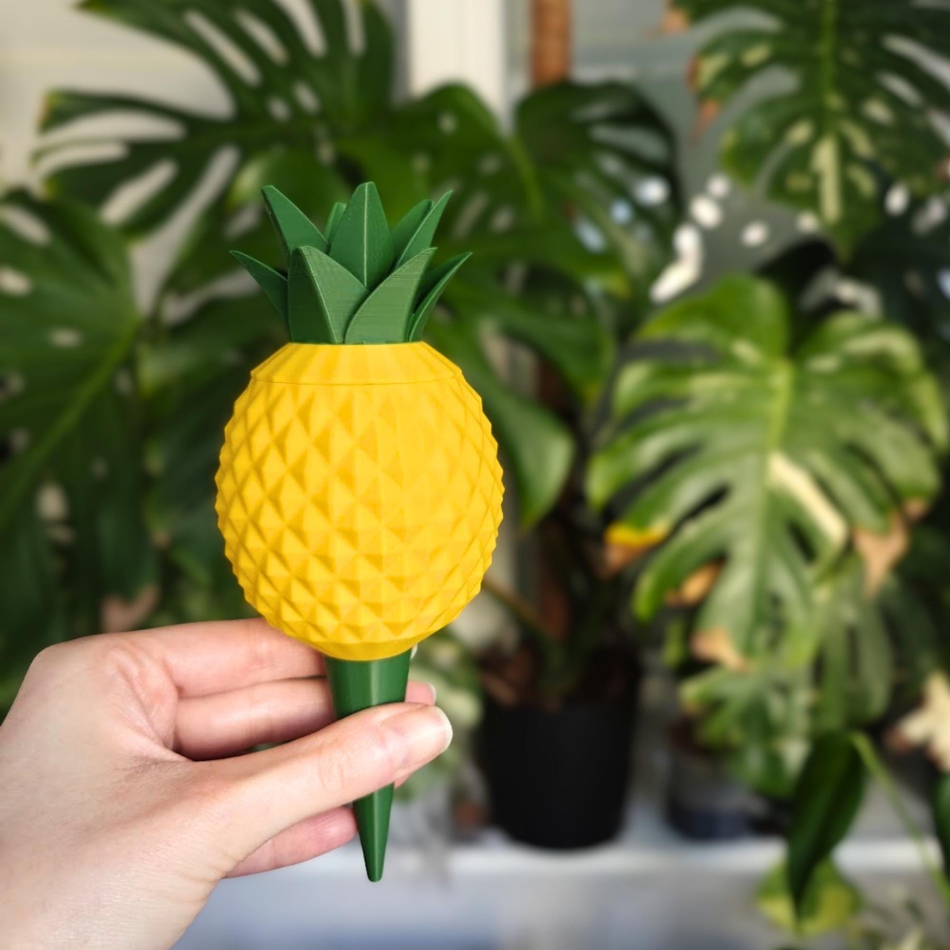D printed pineapple watering spike held in hand in front of indoor houseplants