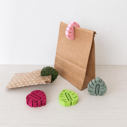 Set of five monstera leaf bag clips in different colors with kraft paper bags on a light tabletop.