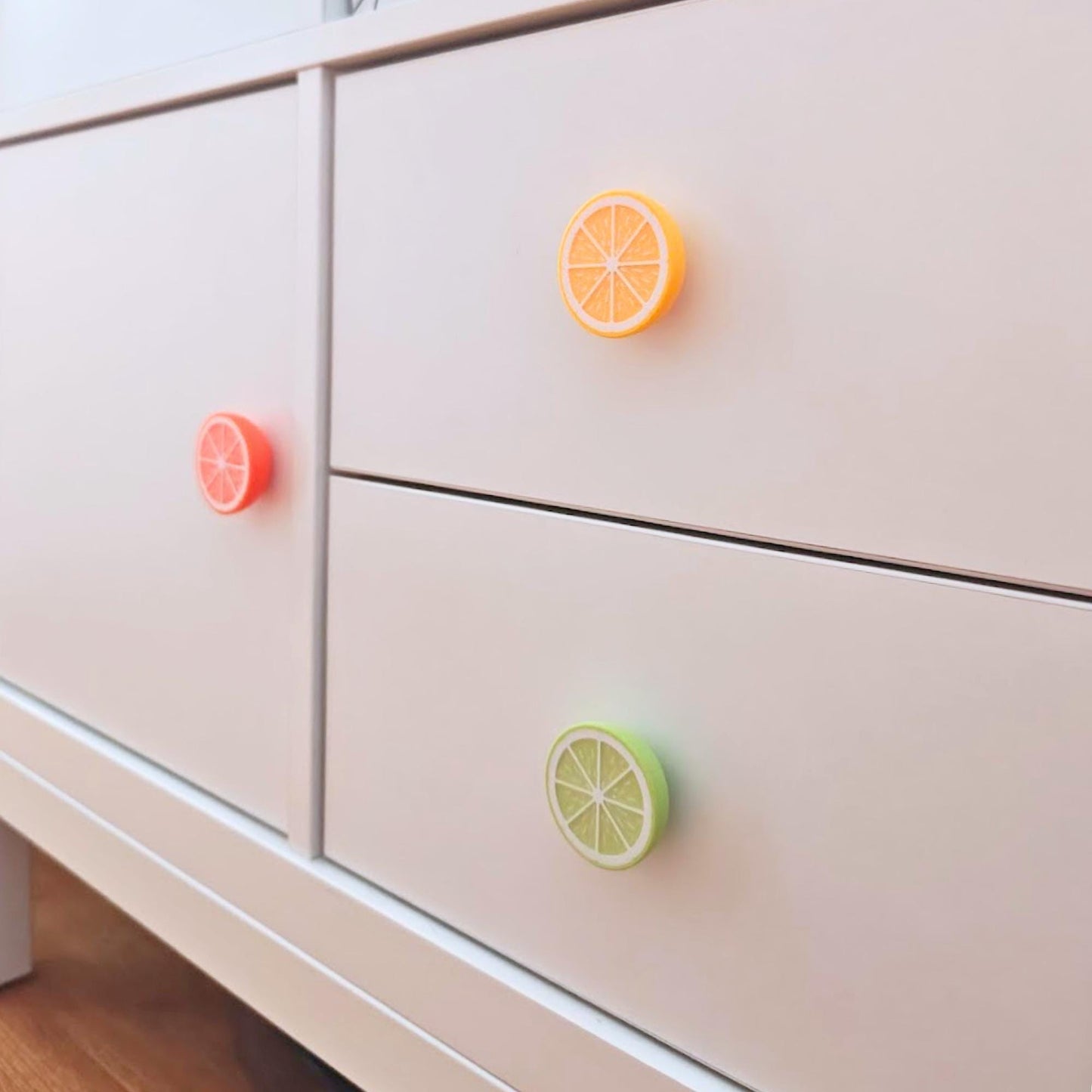 Citrus drawer knobs in lemon, lime, and orange mounted on a white dresser, shown as colorful fruit slice cabinet handles.
