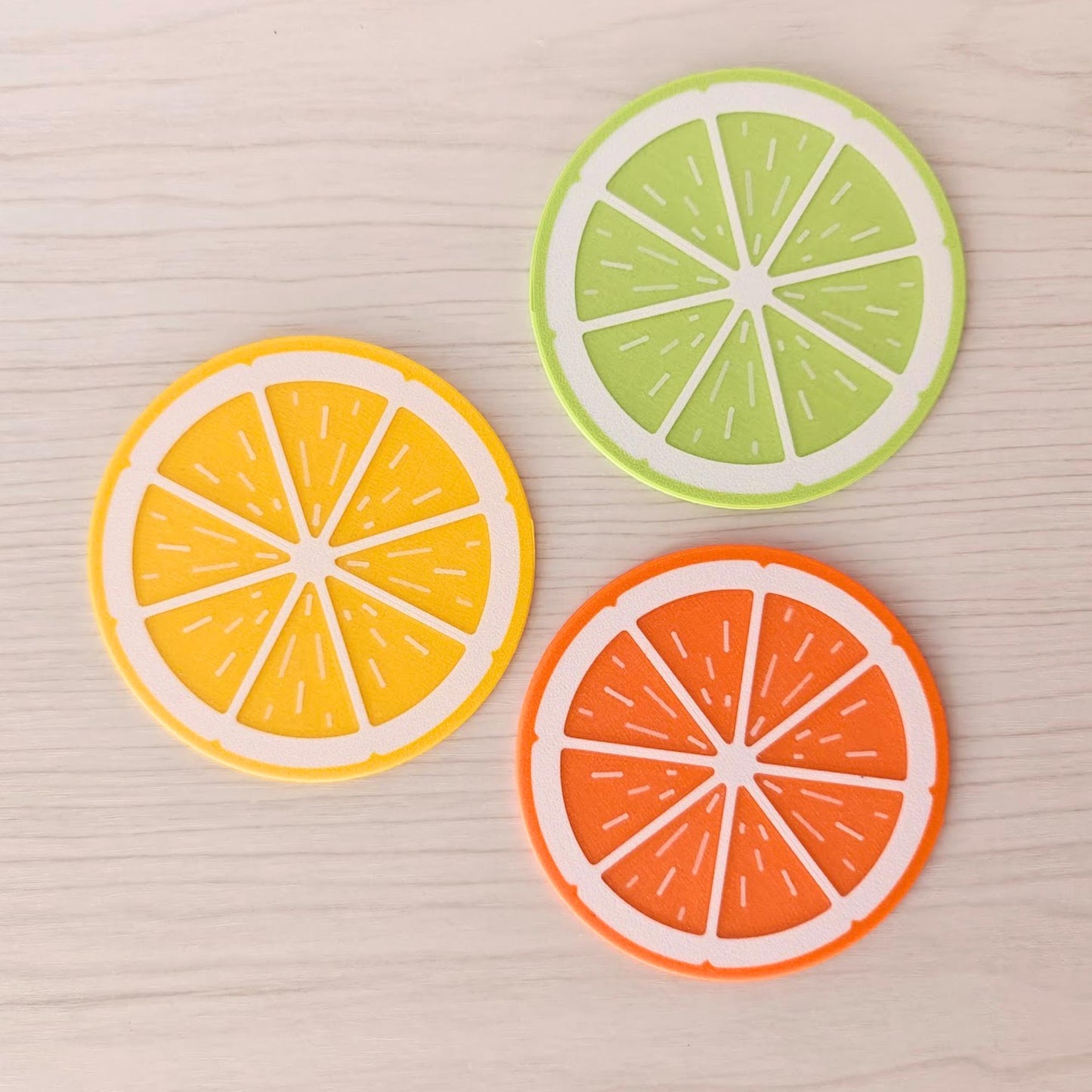Lemon, lime and orange fruit slice coasters arranged on a light wood surface.
