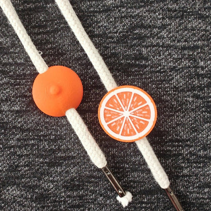 Orange half-fruit drawstring charms on white hoodie cords, close-up view against dark gray fabric.
