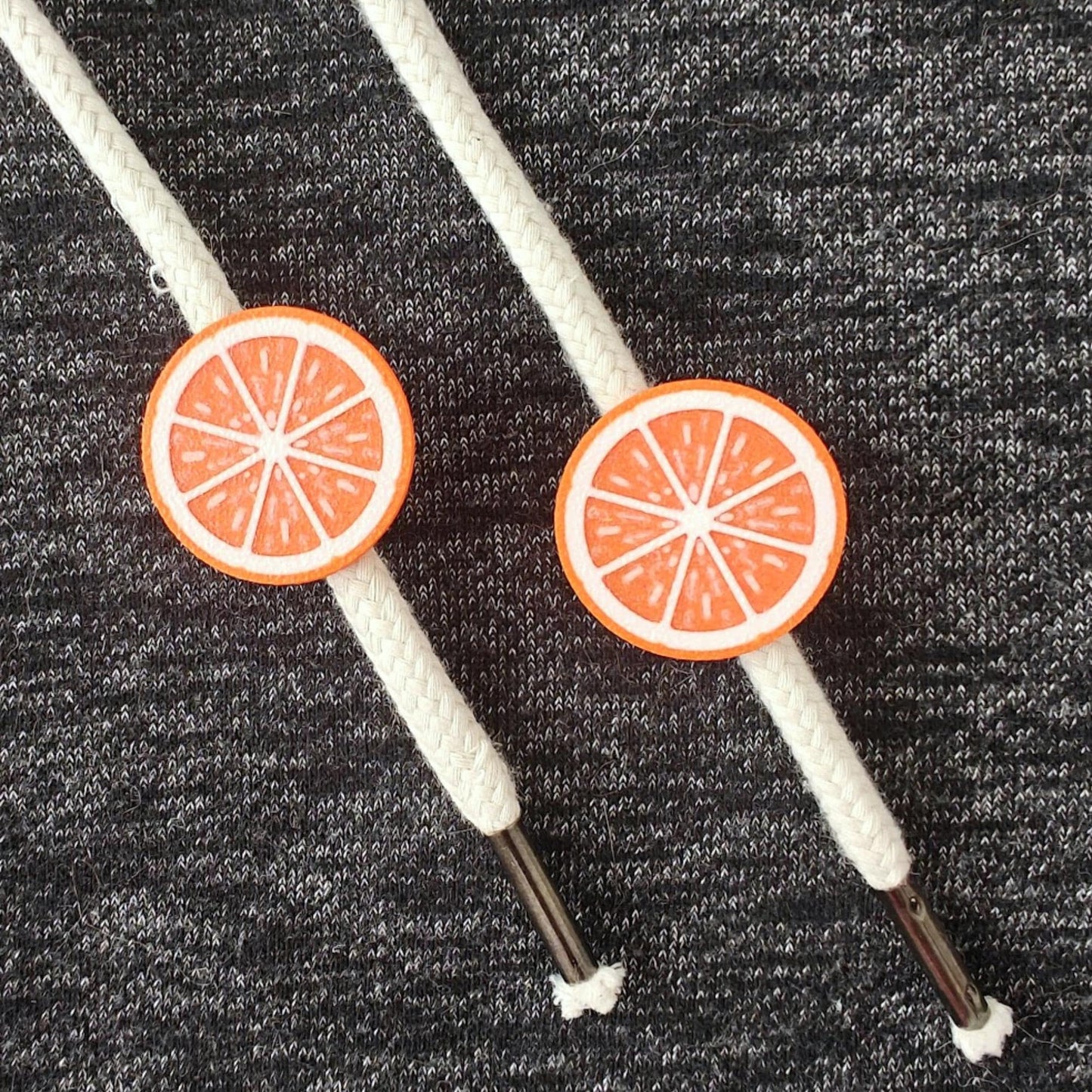 Orange half-fruit drawstring charms on white hoodie cords, close-up view against dark gray fabric.