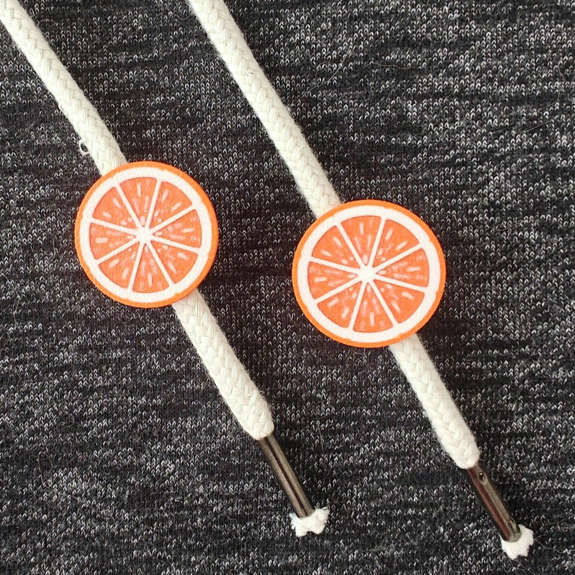 Orange half-fruit drawstring charms on white hoodie cords, close-up view against dark gray fabric.