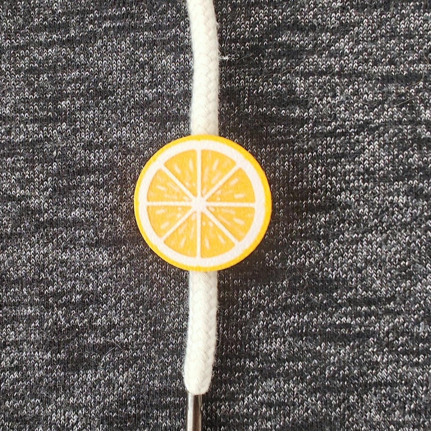 Yellow lemon half-fruit drawstring charm on white hoodie cord, close-up view on dark gray fabric.