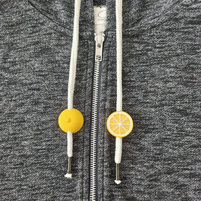 Small yellow citrus drawstring charms on white hoodie cords, shown on dark gray sweatshirt fabric.