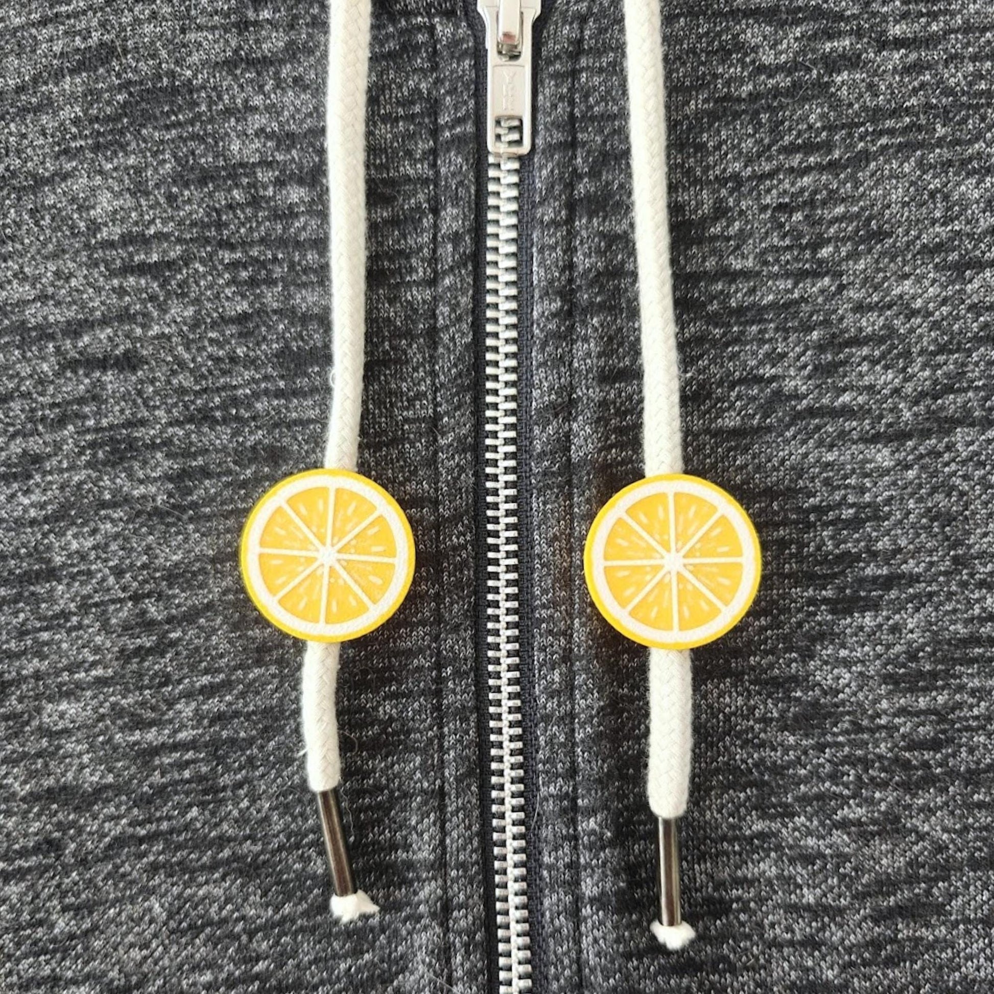 Small yellow citrus drawstring charms on white hoodie cords, shown on dark gray sweatshirt fabric.