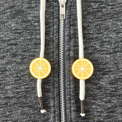 Small yellow citrus drawstring charms on white hoodie cords, shown on dark gray sweatshirt fabric.