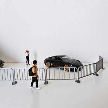 Small silver crowd control barriers arranged in a curved street diorama scene with a car and miniature figure.