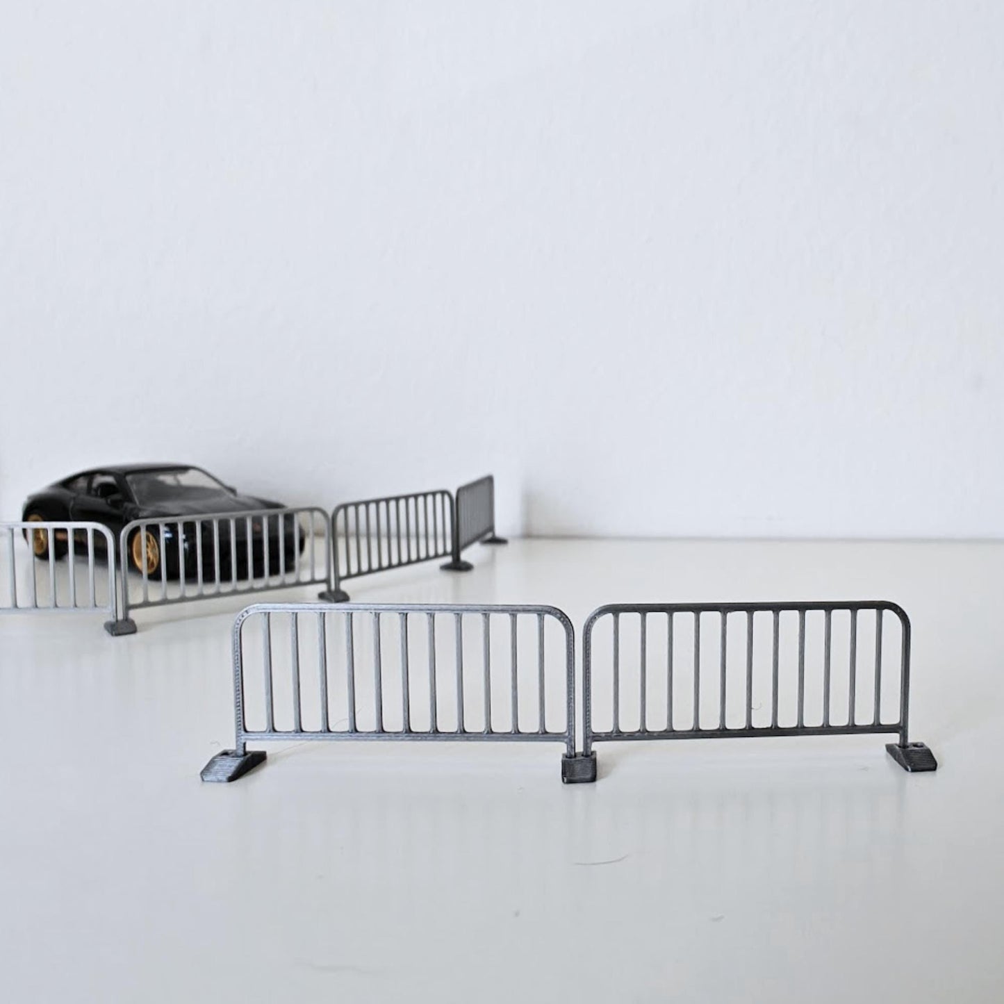Two assembled miniature metal barricades shown from the front with additional barriers and a diecast car in the background.