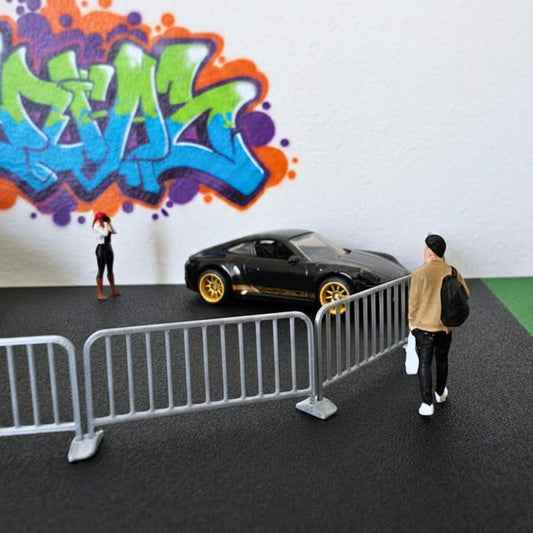 Silver 1:64 scale barricades displayed in a street scene with miniature people, a black car, and a colorful graffiti wall.