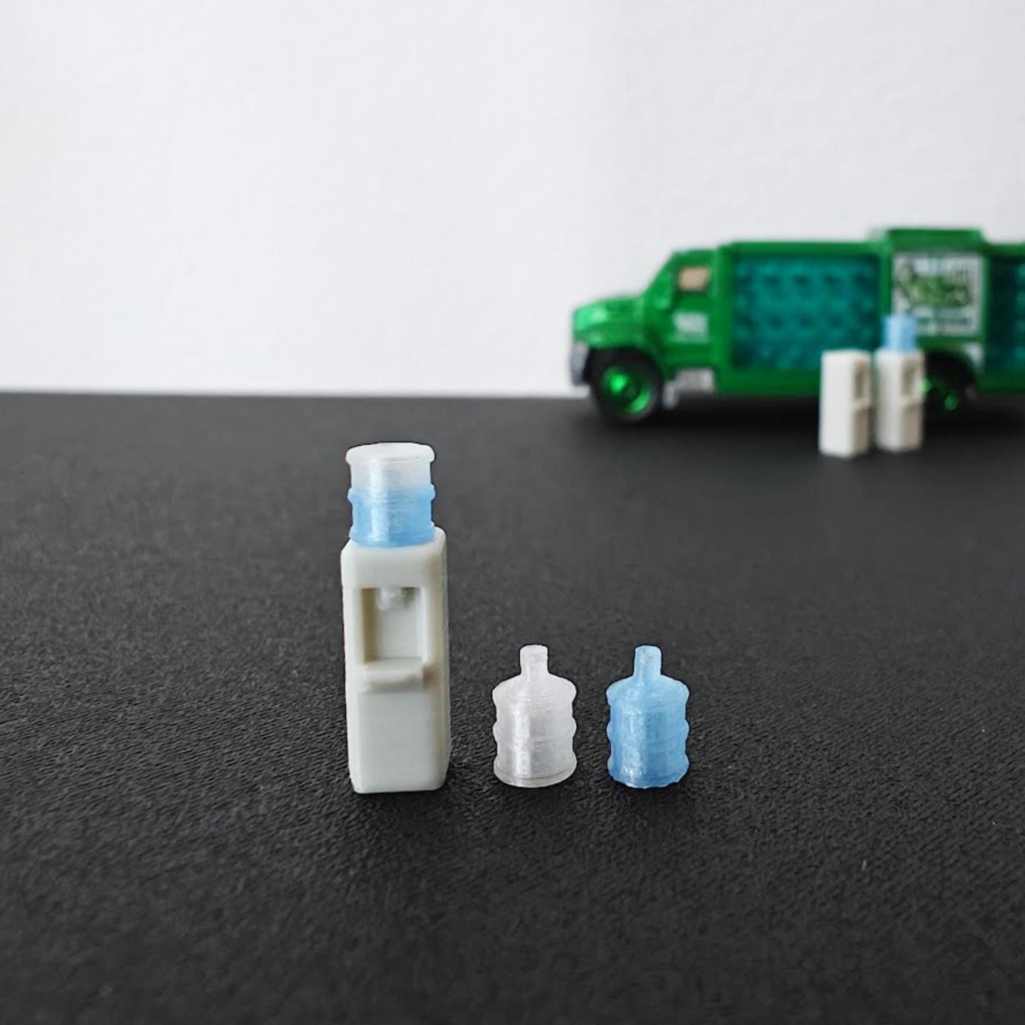 Small 3D printed water dispenser and bottle accessories with a green delivery truck blurred in the background.