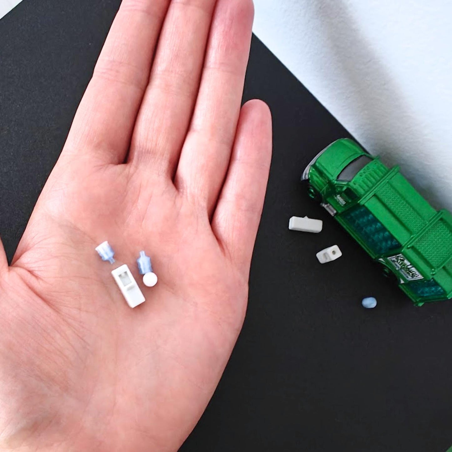 Tiny 1:64 scale water dispenser pieces shown in a hand for size reference next to a green diecast truck.