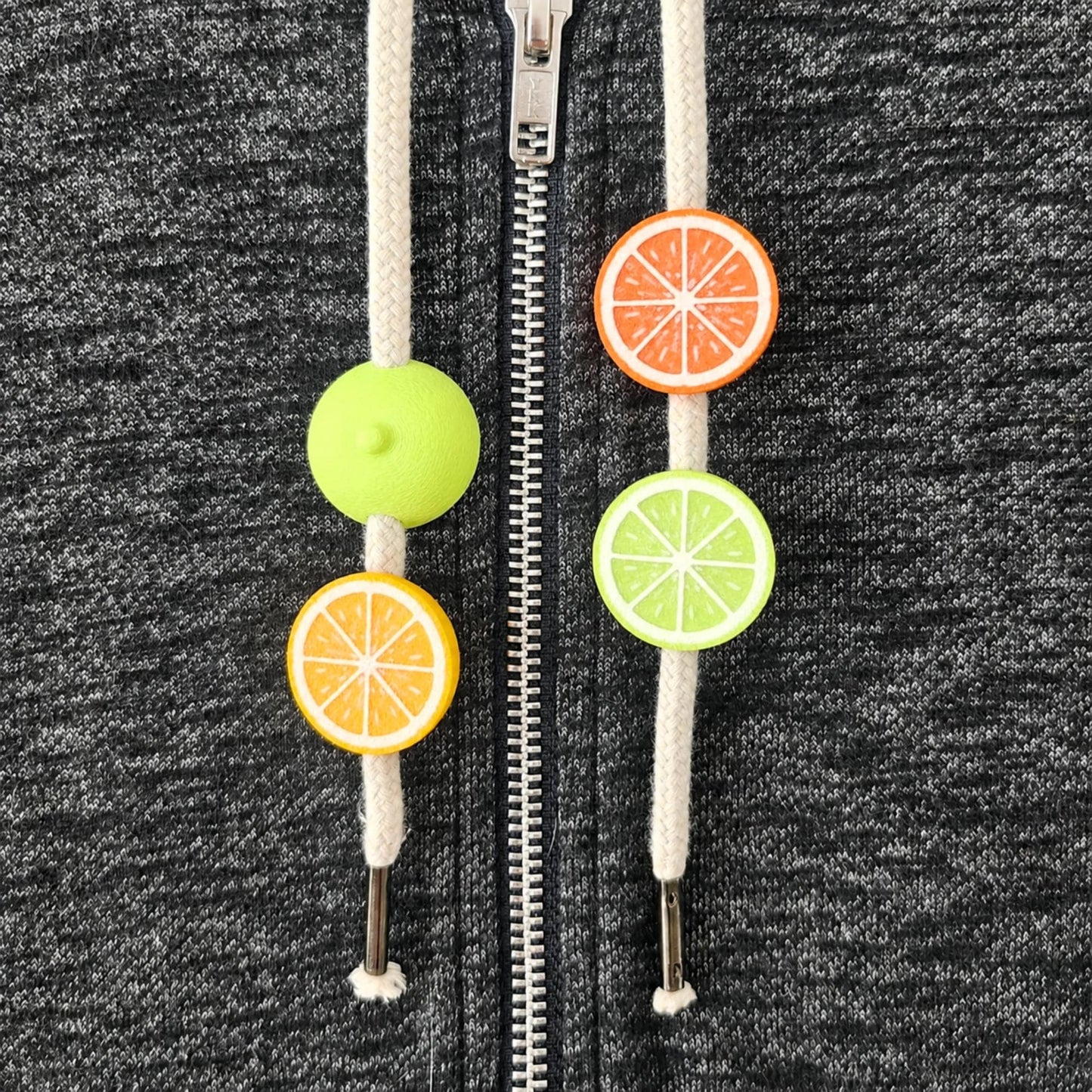 Citrus hoodie drawstring charms with lemon, orange and lime half-fruit beads on white cords against dark gray fabric.