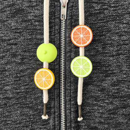 Citrus hoodie drawstring charms with lemon, orange and lime half-fruit beads on white cords against dark gray fabric.