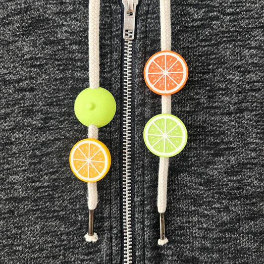 Citrus hoodie drawstring charms with lemon, orange and lime half-fruit beads on white cords against dark gray fabric.
