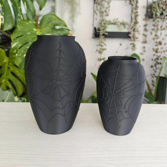 Black spider web vases in two sizes displayed on a light shelf with green plants in the background.