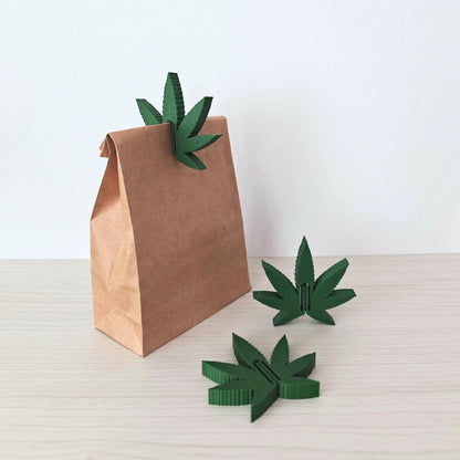 Set of three green cannabis leaf bag clips displayed with a brown paper bag on a light wooden surface.