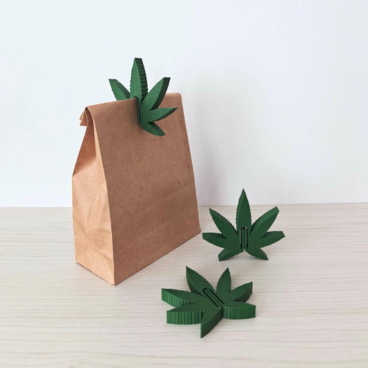 Set of three green cannabis leaf bag clips displayed with a brown paper bag on a light wooden surface.