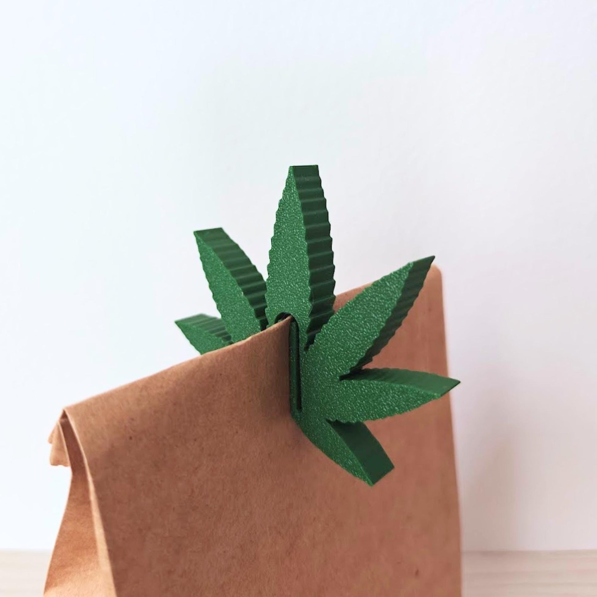 Close-up of a green weed leaf bag clip attached to the folded top of a brown paper bag.