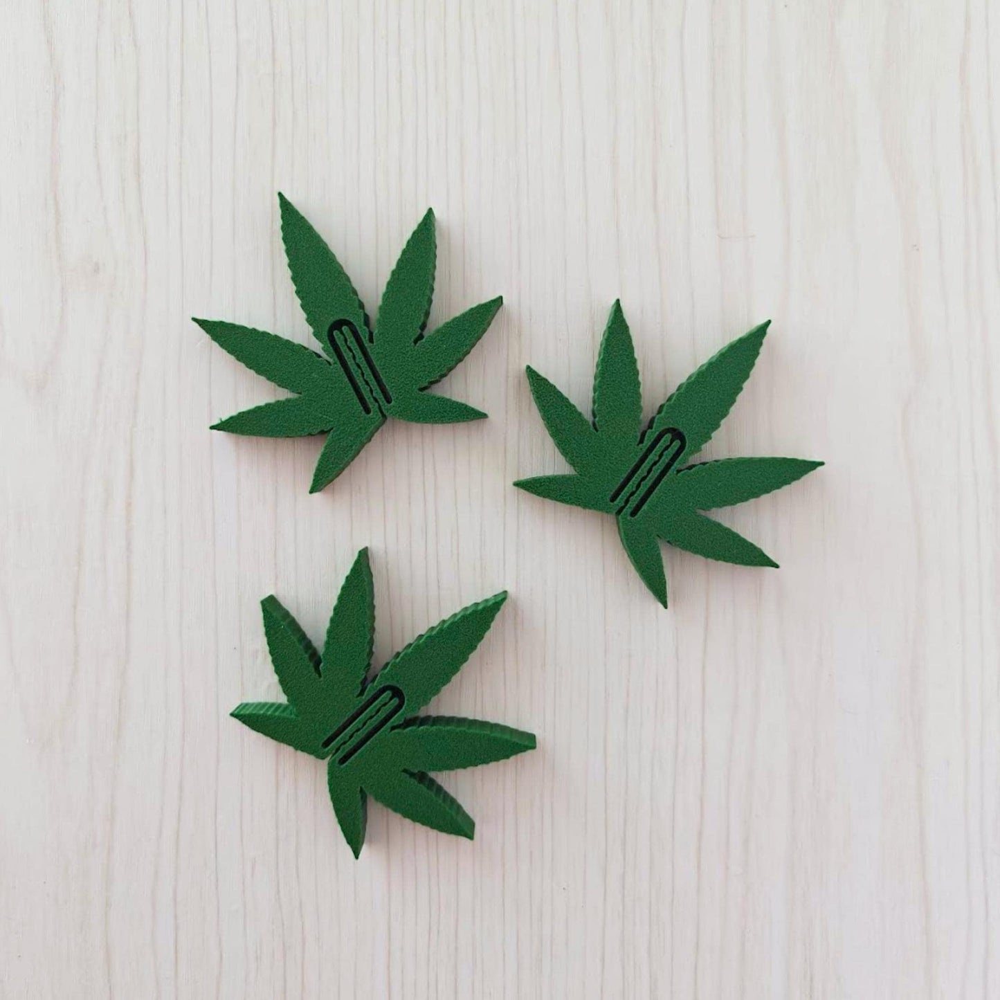 Set of three dark green marijuana leaf snack bag clips arranged on a light wooden surface.