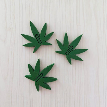 Set of three dark green marijuana leaf snack bag clips arranged on a light wooden surface.