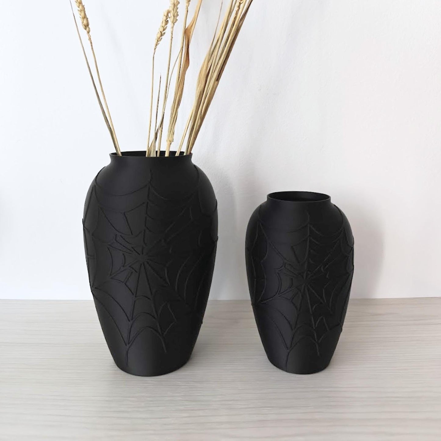 Two black spider web vases displayed with dried flowers on a light wooden shelf.