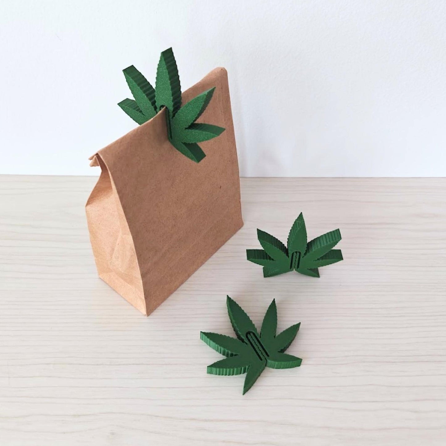 Three marijuana leaf-shaped food clips shown beside a small kraft paper bag against a neutral background.