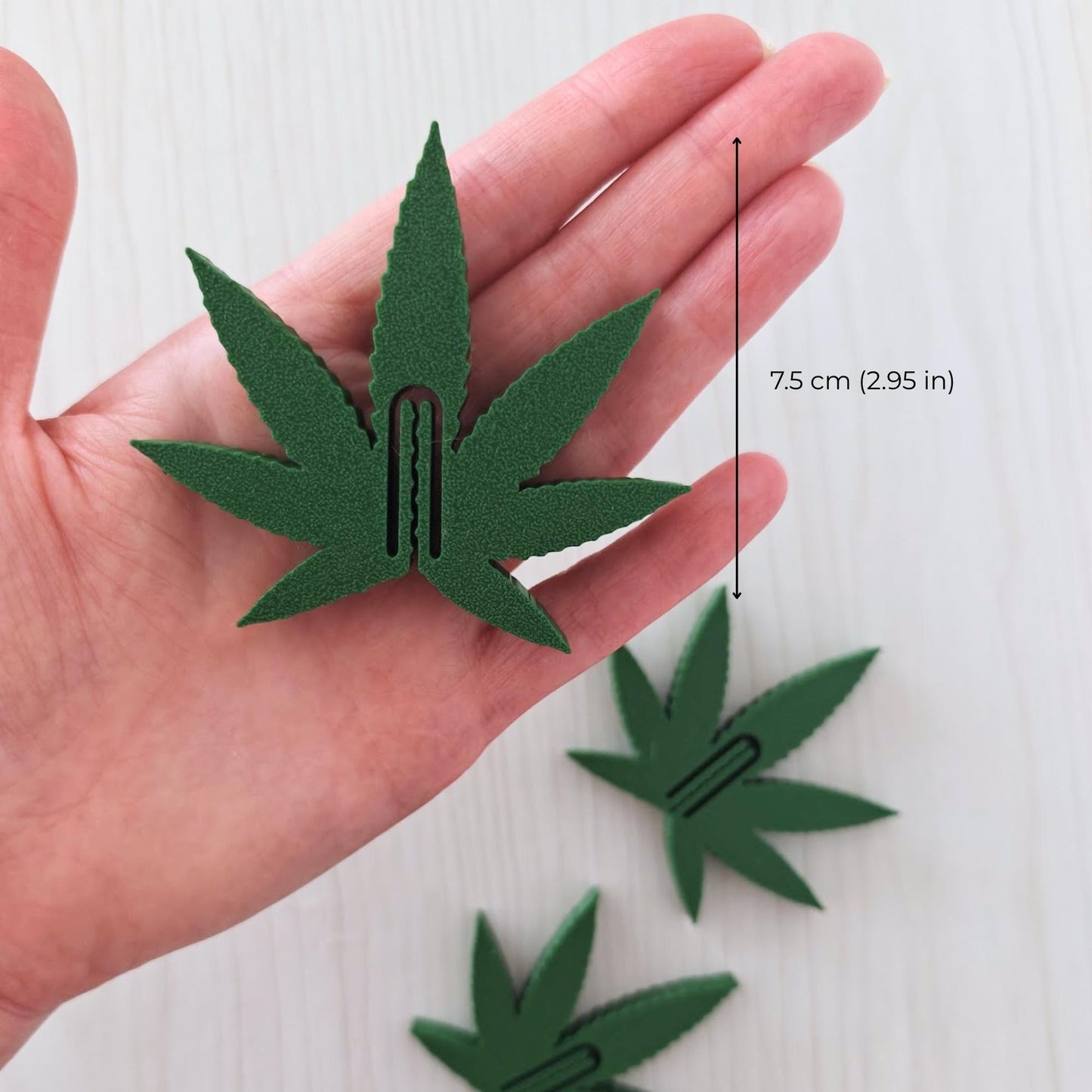 Green cannabis leaf bag clip held in a hand, showing its approximate 7.5 cm (2.95 in) size.