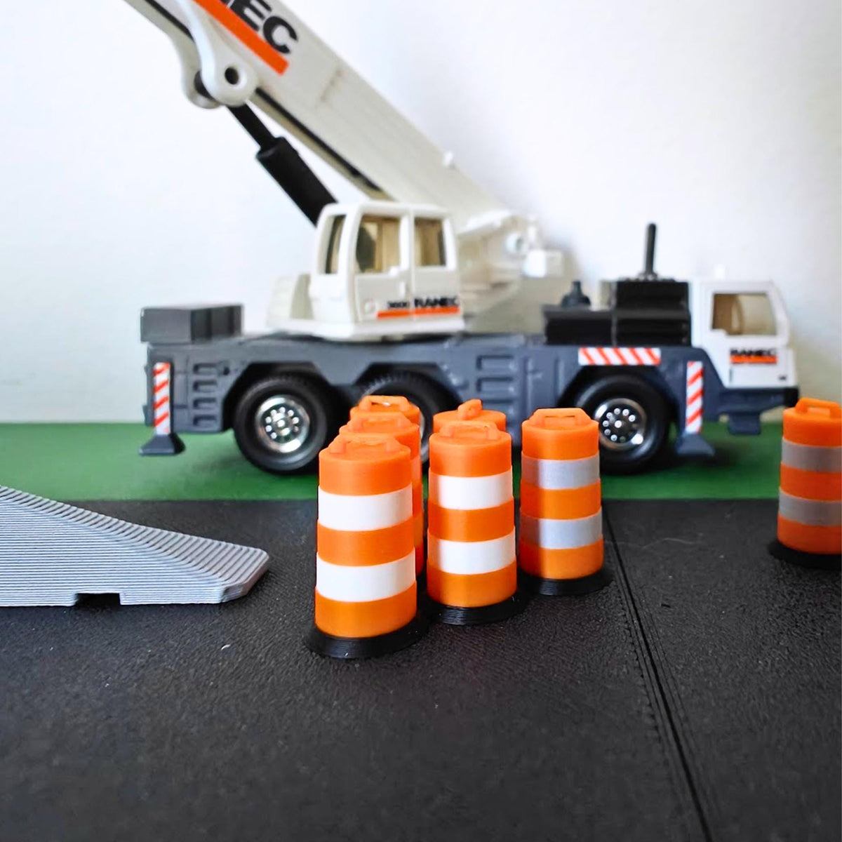 Close-up comparison of miniature traffic barrels in different scales, with orange bodies, silver bands and black bases.