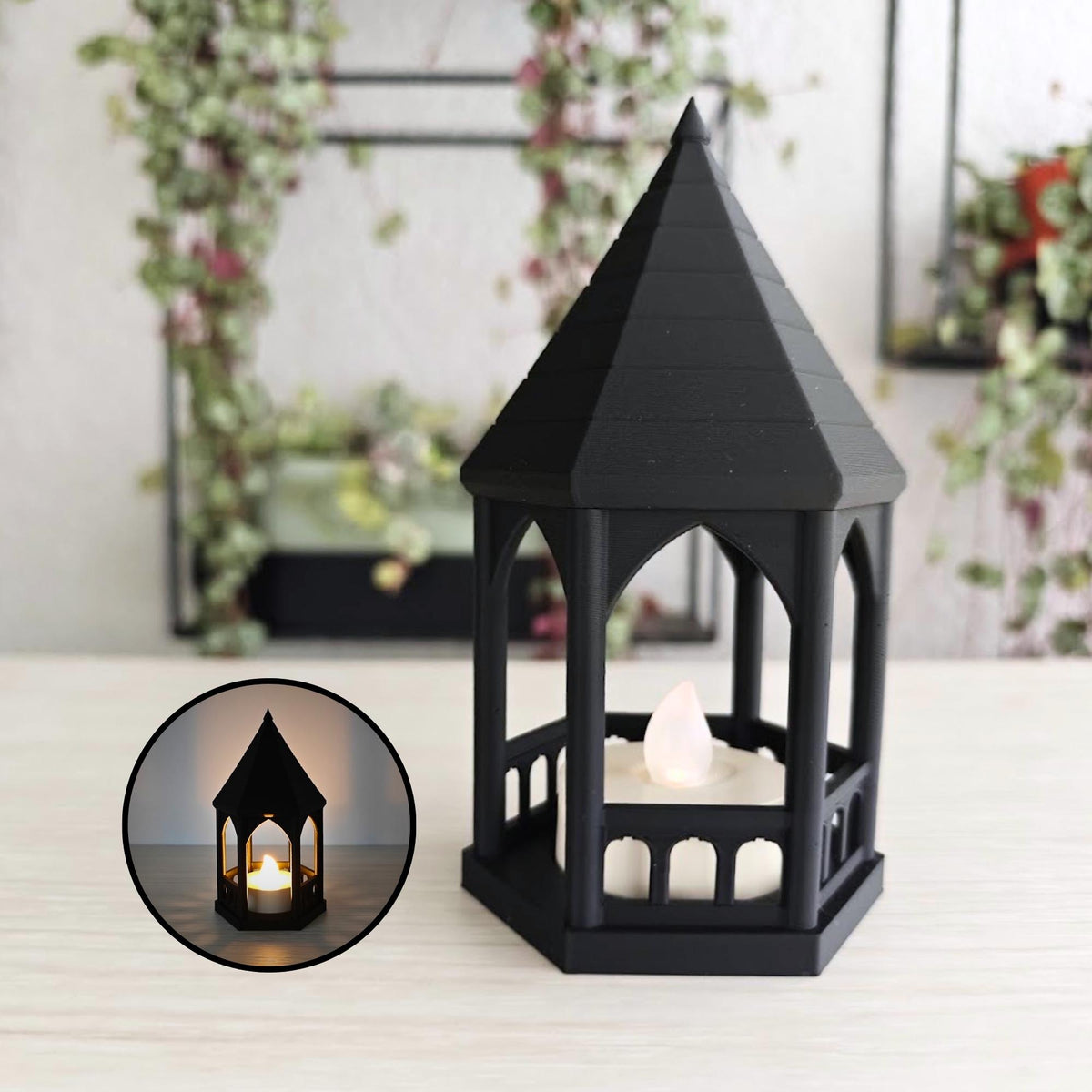A miniature matte black, gothic-style gazebo tealight holder with a lit LED candle inside, sitting on a light wooden desk with a hanging plant blurred in the background.