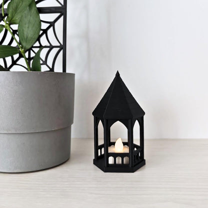 The black gazebo tealight holder with a lit LED candle, placed next to a grey potted houseplant with a black trellis.