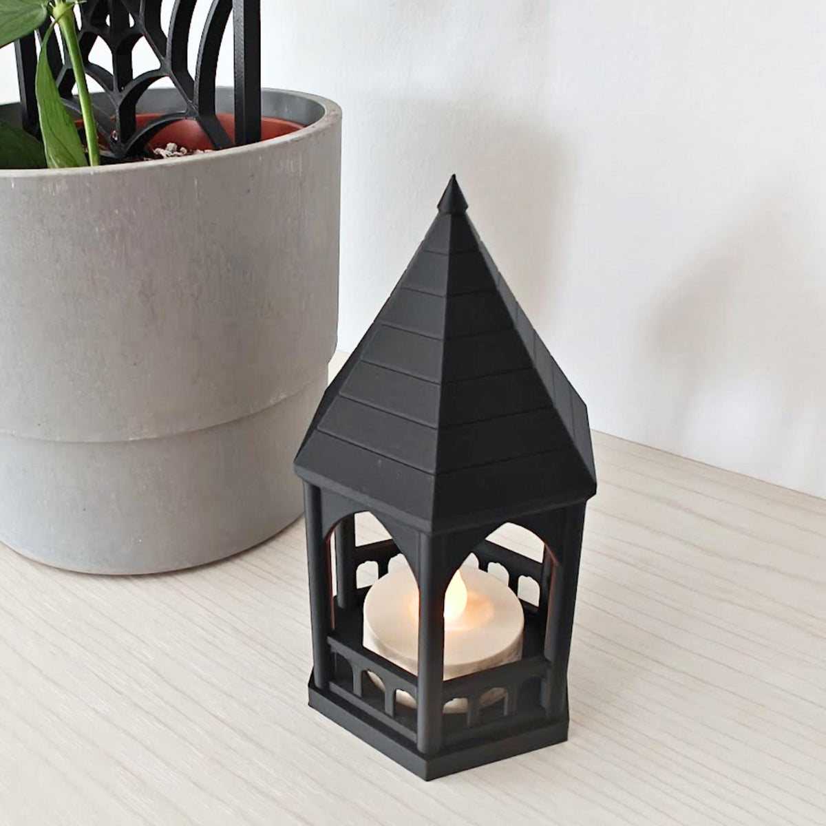A high-angle view of the black gazebo tealight holder glowing with an LED candle, sitting next to a potted plant.