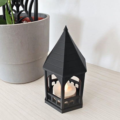 A high-angle view of the black gazebo tealight holder glowing with an LED candle, sitting next to a potted plant.