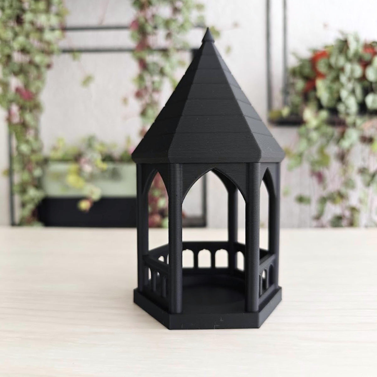 Angled View: A slightly angled shot of the black gothic gazebo tealight holder featuring an LED candle inside, with a blurred plant in the background.