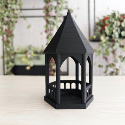 Angled View: A slightly angled shot of the black gothic gazebo tealight holder featuring an LED candle inside, with a blurred plant in the background.