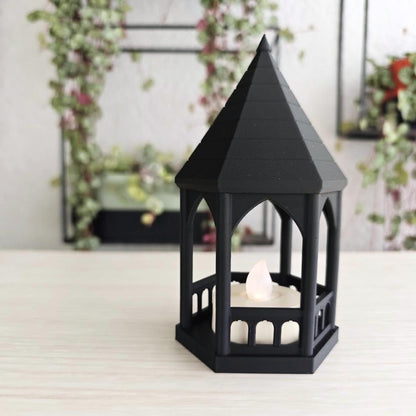 A miniature matte black, gothic-style gazebo tealight holder with a lit LED candle inside, sitting on a light wooden desk with a hanging plant blurred in the background.