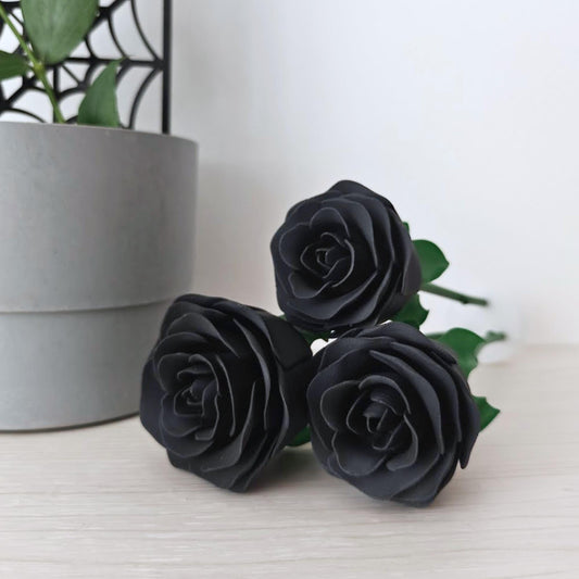 Set of three matte black faux roses displayed upright on a light wooden surface beside a grey plant pot.
